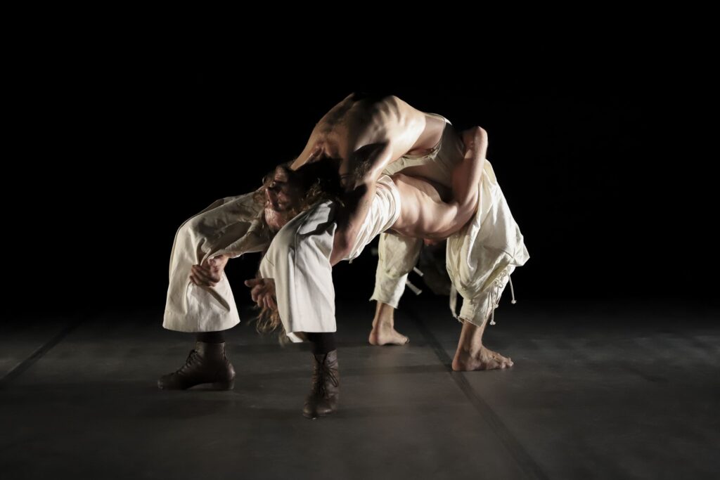 Dos bailarines flexionados uno sobre otro haciendo un arco sobre un escenario con fondo negro