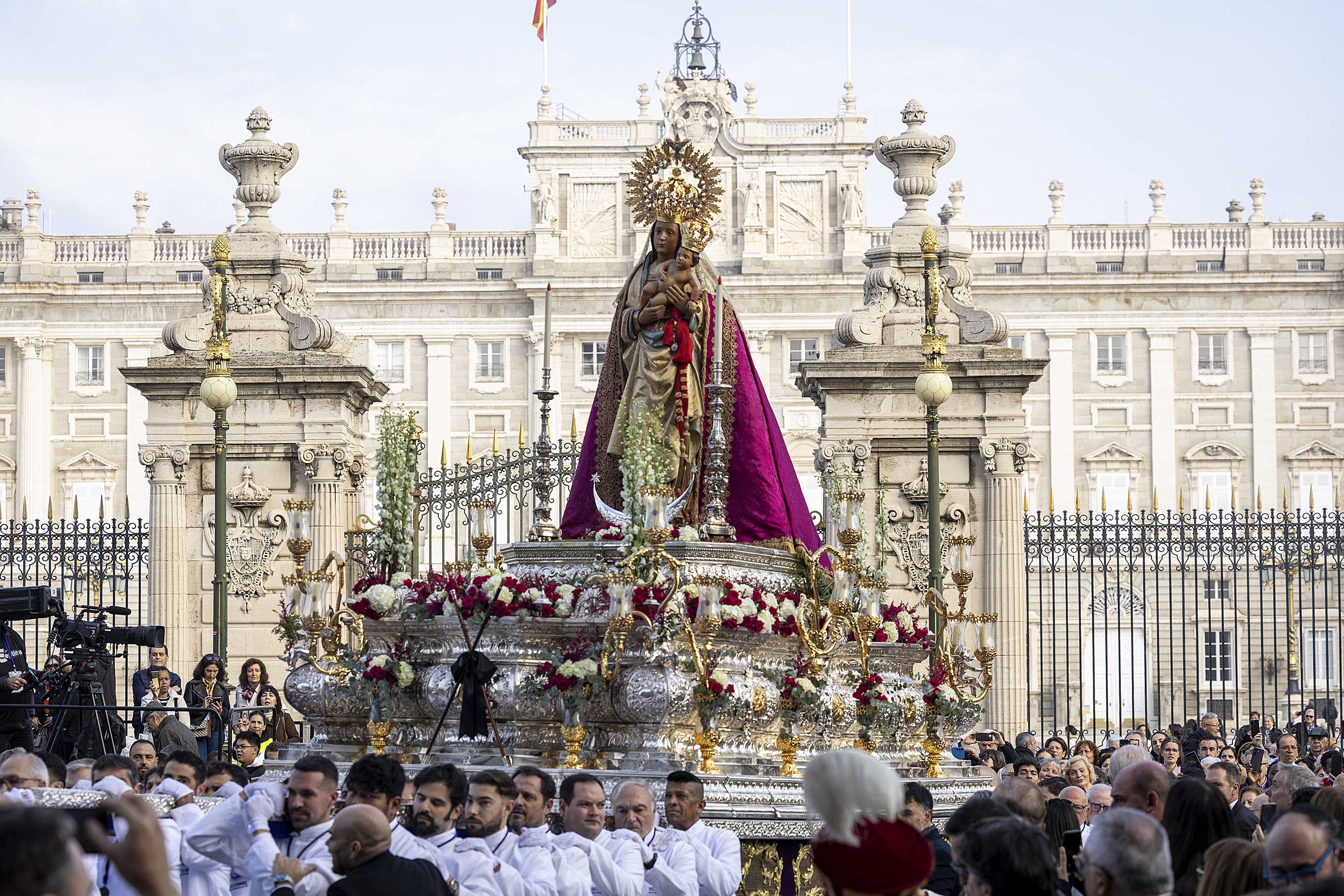 Procesión religiosa en Madrid con la imagen de la Virgen de la Almudena, vestida con manto púrpura y dorado, portada por costaleros frente al Palacio Real, rodeada de flores y numerosos asistentes. Uno de los actos más destacados dentro de la agenda cultural del puente de la Almudena