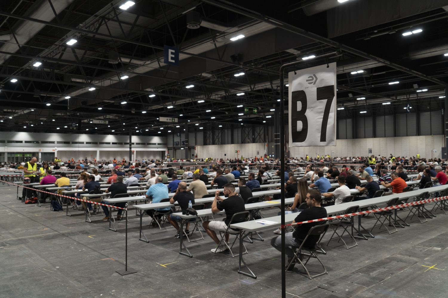Esta imagen captura un momento de alta exigencia donde una multitud de aspirantes a conductores se congregan en un inmenso centro de convenciones para realizar un examen oficial de la EMT. Los candidatos aparecen sentados en filas interminables de mesas individuales, reflejando el proceso de selección masivo y la organización logística necesaria para evaluar a tantos participantes a la vez. El entorno industrial y la señalización técnica subrayan la seriedad de la convocatoria laboral, diseñada para filtrar a los profesionales que formarán parte del sistema de transporte público.